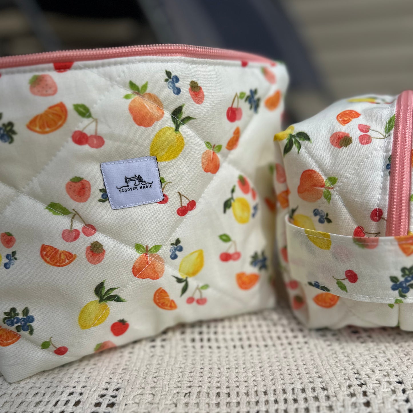 Two quilted pouches with fruit pattern and visible brand label on a textured surface.